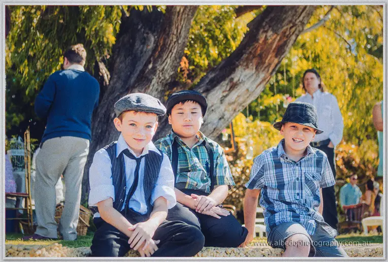 Three boys in flat caps, braces and check shirts sit grinning under a sprawling willow at the picnic