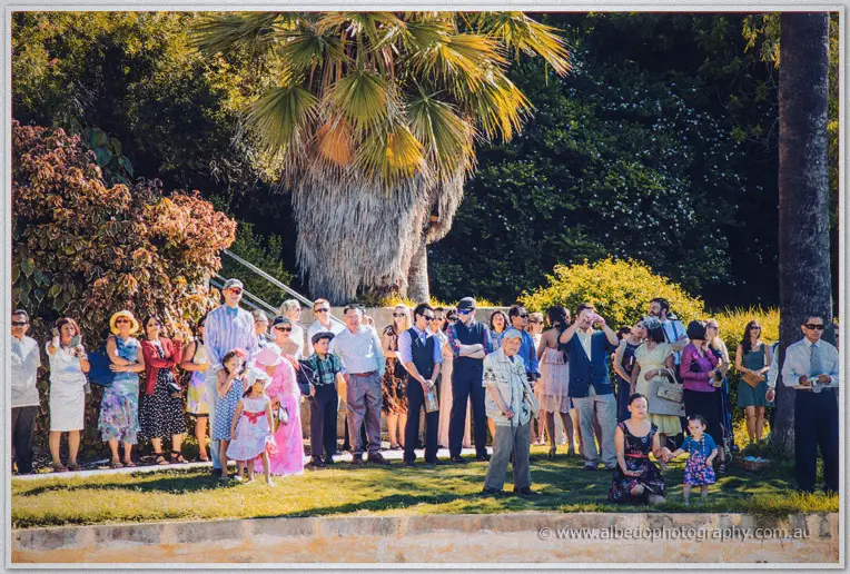 Crowds of guests and children line the sunlit lawn under palms awaiting the ceremony