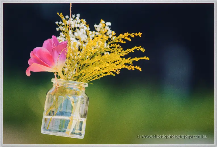 A suspended glass jar holds pink freesia, wattle and baby's breath against soft green bokeh