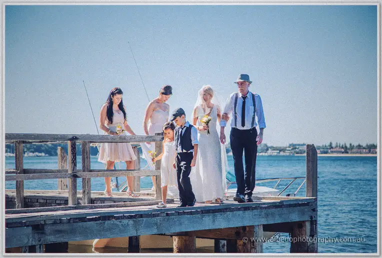 Tansy arrives on the weathered jetty arm in arm with her father, party trailing behind