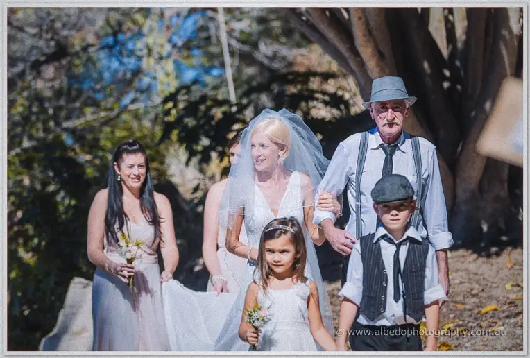 Tansy walks the bushland path veiled beside her father with bridesmaids and children following