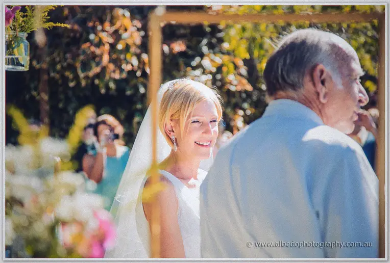 Tansy smiles past her father's shoulder as guests photograph her arrival beneath the tree canopy