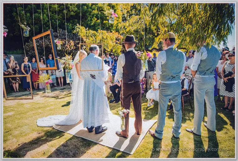 Tansy faces the priest under hanging frames and jars while groomsmen in braces stand by
