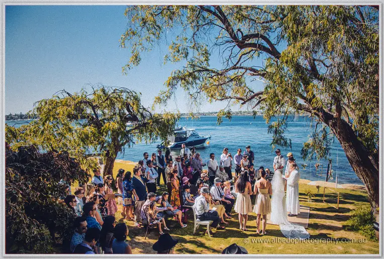 Wide view of guests gathered on the riverbank lawn as Tansy and Ciaran marry under the trees