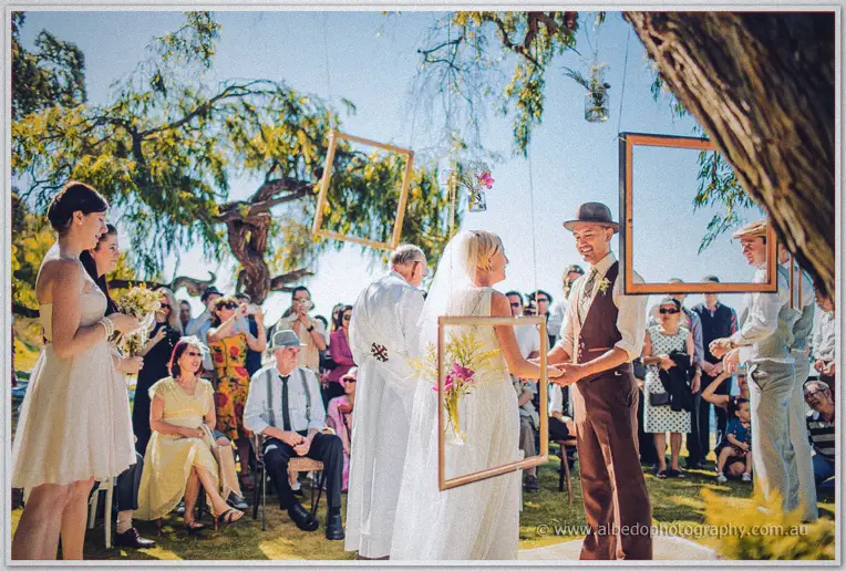 Tansy and Ciaran clasp hands before the celebrant framed by willows and hanging frames