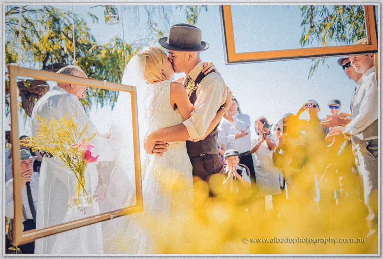 Tansy and Ciaran kiss beneath hanging frames as yellow wattle blurs across the foreground