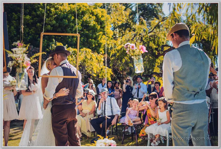 Tansy and Ciaran embrace framed by a hanging frame as smiling guests look on in dappled sun