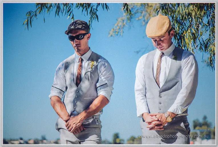 Two groomsmen in flat caps and grey vests stand solemnly under willow branches awaiting the bride