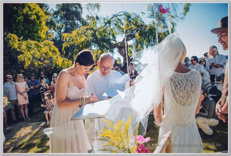 A bridesmaid signs the marriage register beside the priest as Tansy waits veiled among guests