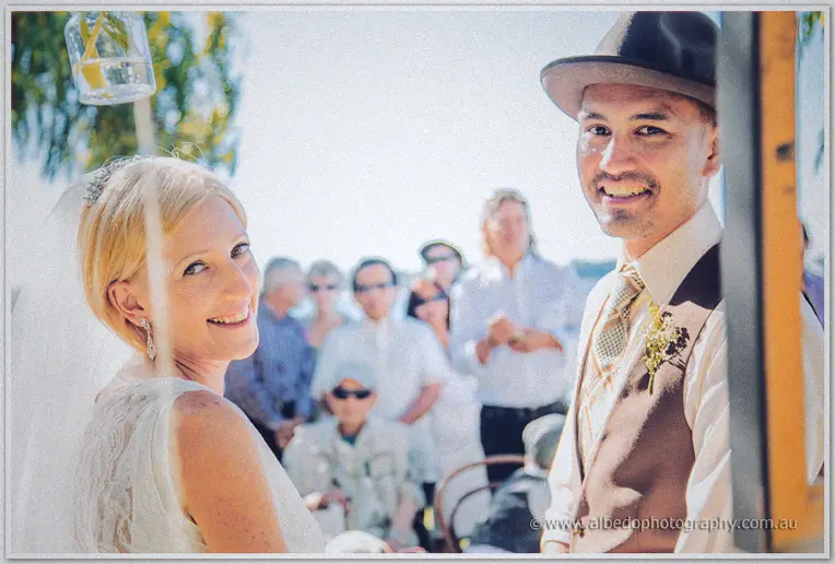 Tansy and Ciaran turn beaming toward the camera amid their guests in bright riverside sunshine