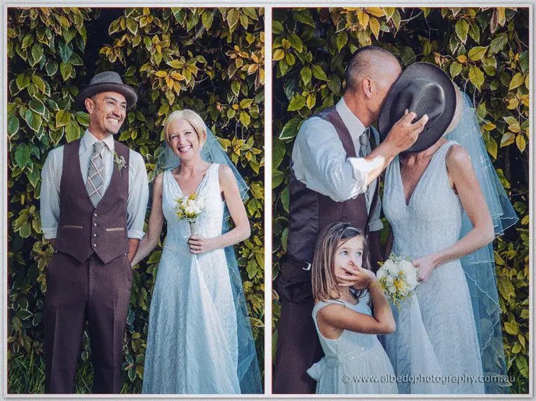 Groom in a flat cap kisses his veiled bride under green leaves while their flower girl giggles