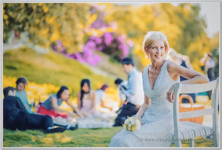 Bride Tansy smiles from a white chair holding yellow posies while guests picnic behind her