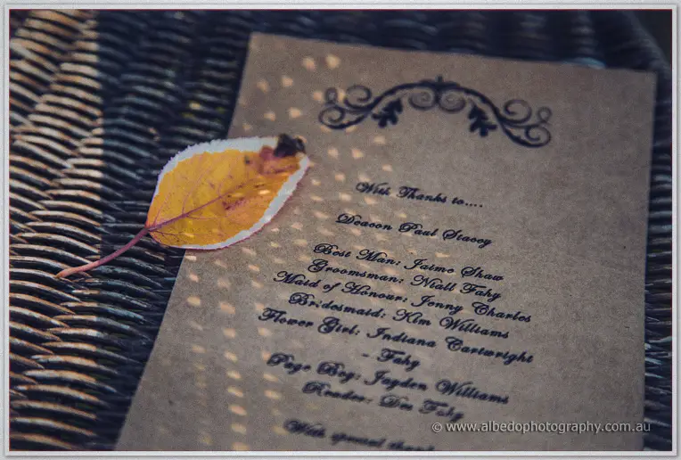 A kraft paper card thanking the wedding party rests on wicker beside a single autumn leaf