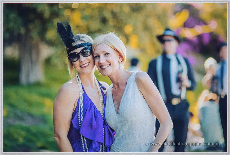 The bride laughs with a guest in a purple flapper dress and feathered headband in golden light