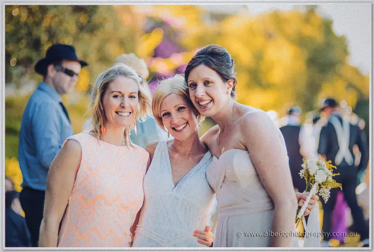 Three smiling women in pastel dresses hug during the sunny picnic wedding at Mosman Park