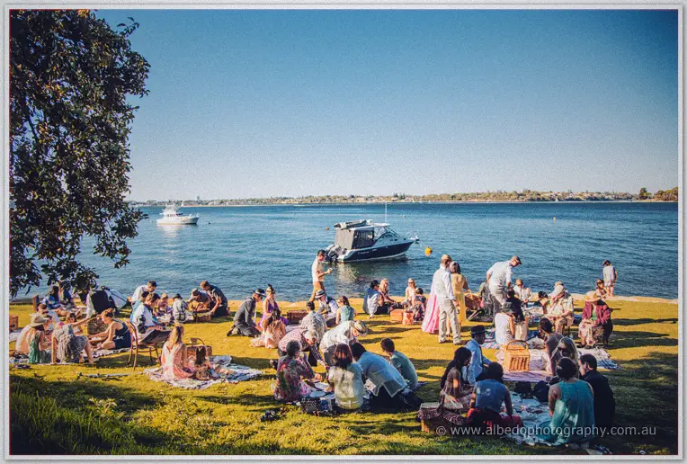 Wedding guests picnic on rugs by the blue river as boats float past Green Place Reserve