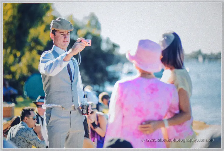 A groomsman in a flat cap snaps photos by the river as guests enjoy the picnic celebration