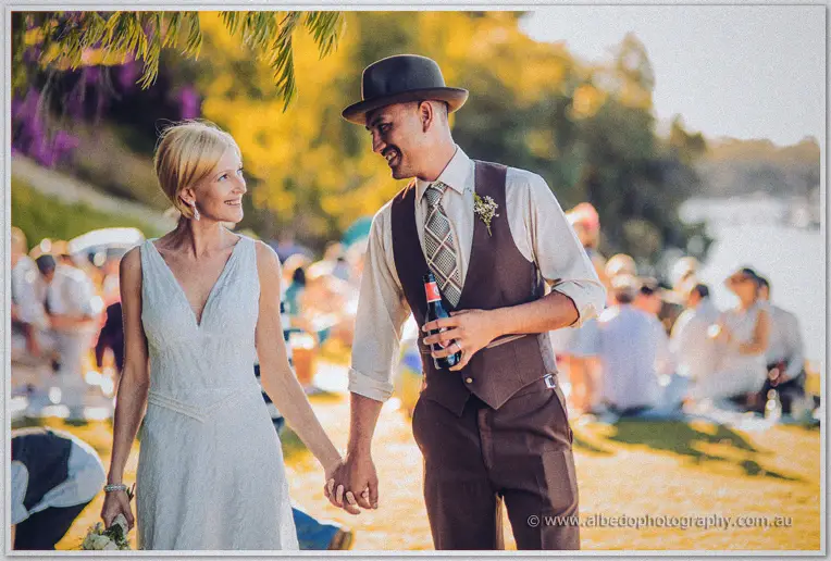 The newlyweds stroll hand in hand past picnic rugs in warm afternoon light