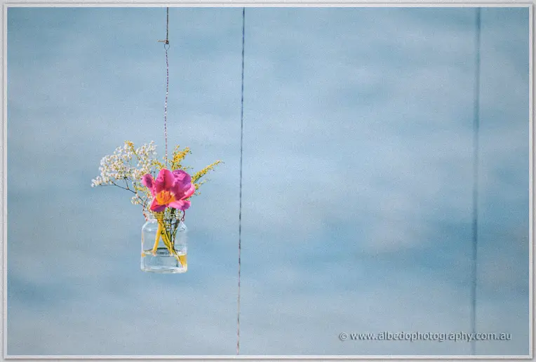 A glass jar of pink blooms and baby's breath hangs on a string against calm blue river water