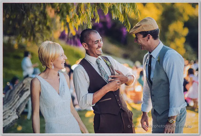 The groom shares a laugh with his groomsman and the bride under golden trees