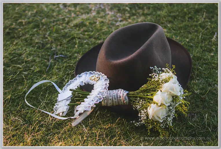 A felt trilby hat rests on the grass with a white rose boutonniere and ribbon-wrapped bouquet