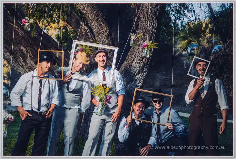 Groomsmen in suspenders hold vintage frames around their faces beneath a flowering gum tree