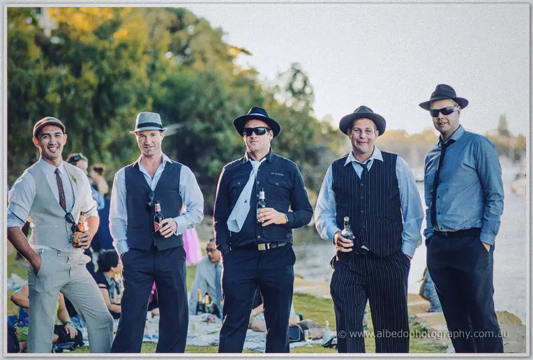 Five men in trilby hats and waistcoats pose with beers along the Swan River foreshore