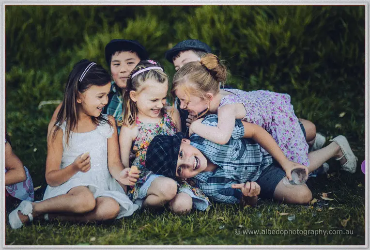 Children in flat caps pile giggling on the grass during the vintage picnic wedding