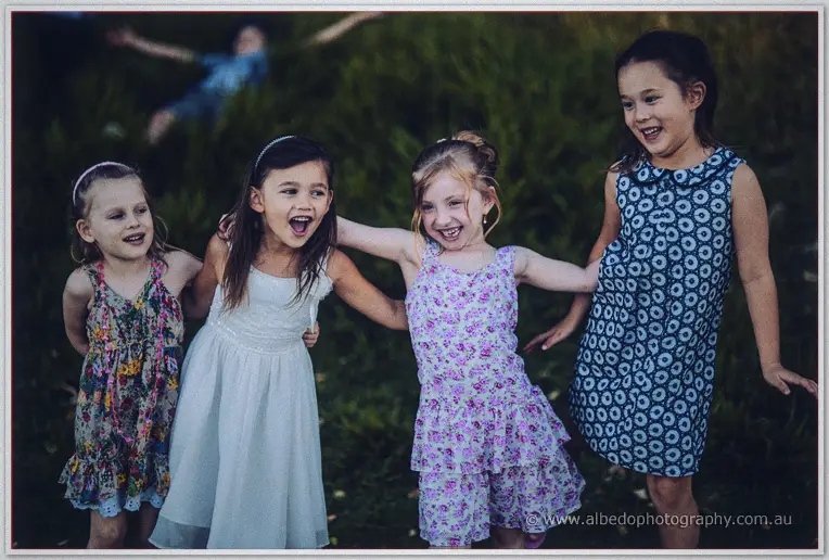 Four girls in summer dresses link arms and laugh on the lawn in the last light