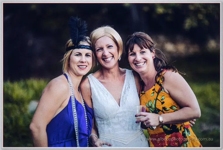The bride beams between a friend in flapper pearls and another in bright florals at dusk