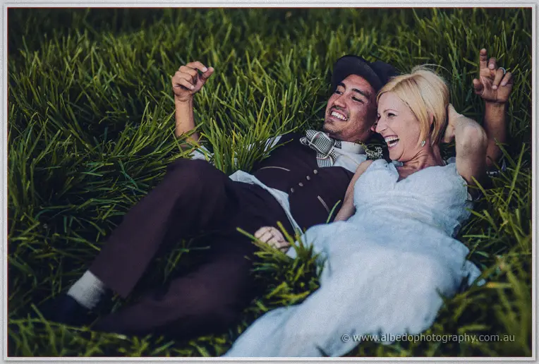 The bride and groom sprawl in long grass laughing with arms flung wide at dusk