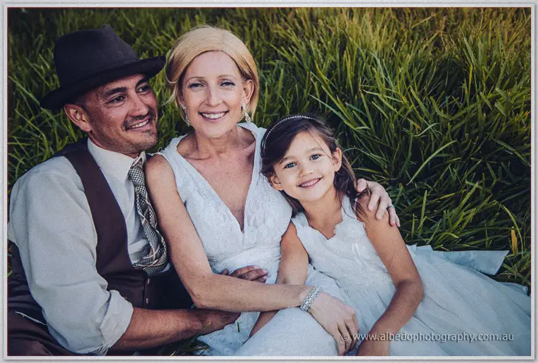 The newlyweds sit in the grass with a young flower girl snuggled between them