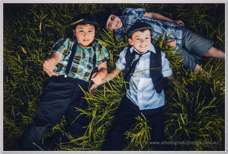 Three boys in flat caps and suspenders grin up from the long grass below the camera