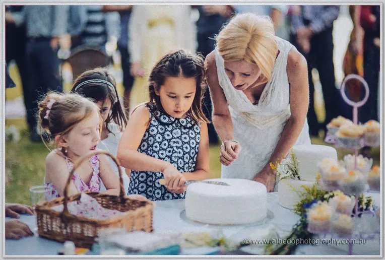 Tansy helps a young guest cut the white wedding cake as girls watch at the picnic table