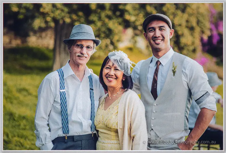 An older couple in suspenders and a feathered fascinator pose smiling beside the groom