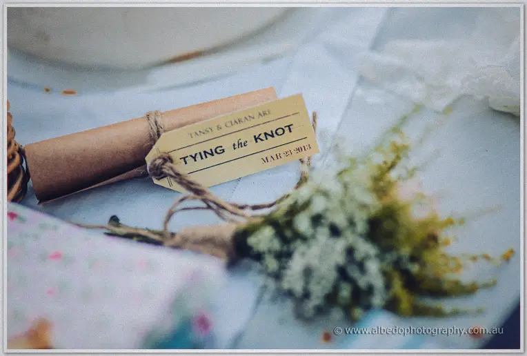 A kraft scroll tag reading Tying the Knot rests beside a baby's breath sprig on a blue table