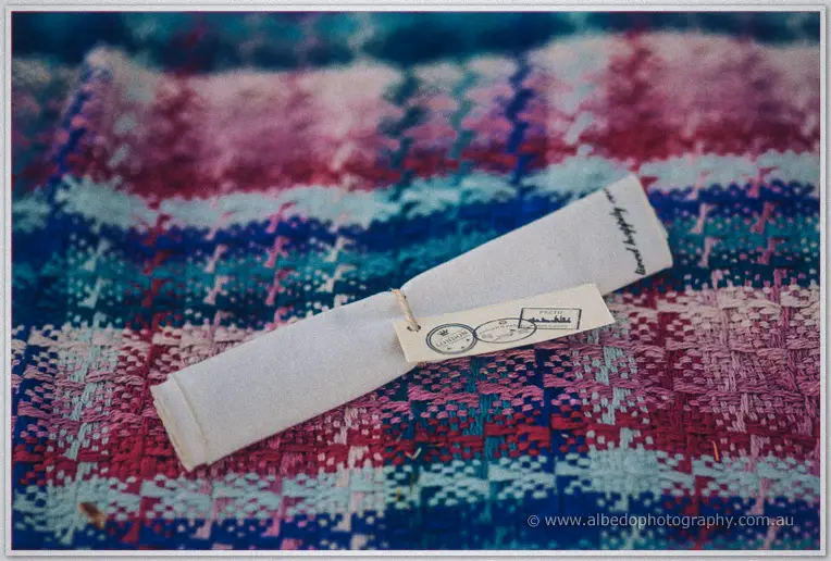 A rolled guest favour with a vintage postage tag rests on a colourful woven picnic rug