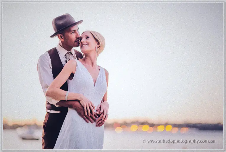 Ciaran in a trilby kisses Tansy's temple as city lights blur across the river behind them