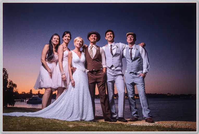 Tansy, Ciaran and their wedding party stand arm in arm by the Swan River under a dusk sky