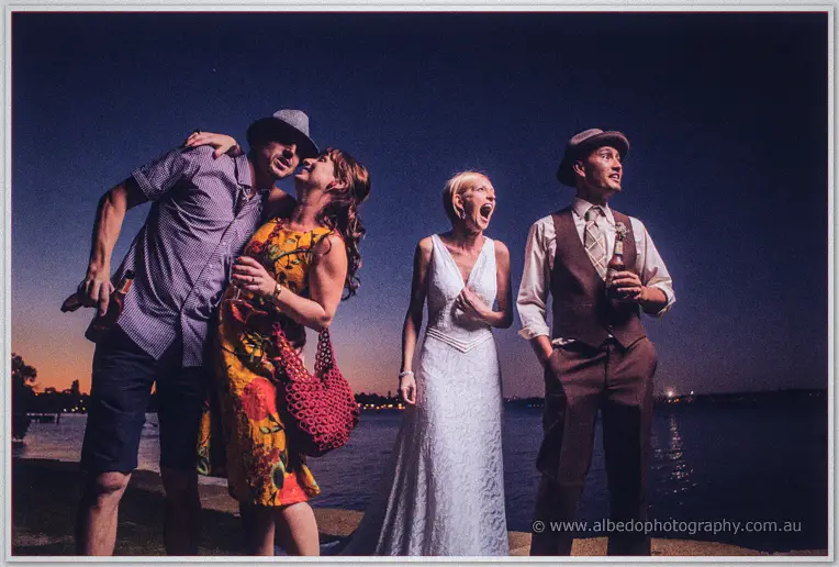 Friends pose goofily around the laughing newlyweds on the riverbank after dark