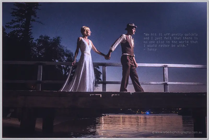 Tansy and Ciaran walk hand in hand along a timber jetty over calm water beneath a deep blue dusk
