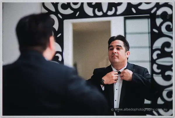 Groom studies his reflection while fastening his black bow tie in an ornate framed mirror