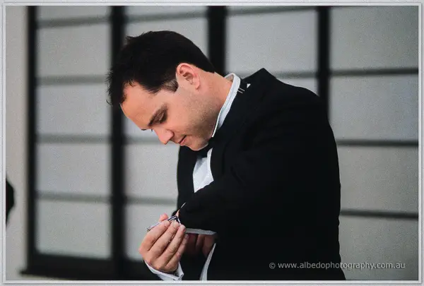 Groom's man in a dark suit fastens his cufflink while looking down beside a lattice window