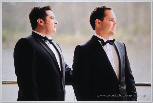 Two groomsmen in matching black tuxedos stand shoulder to shoulder before a misty lake backdrop
