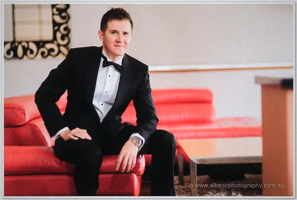 Groom in bow tie and tuxedo perches on a red leather couch arm glancing at his watch
