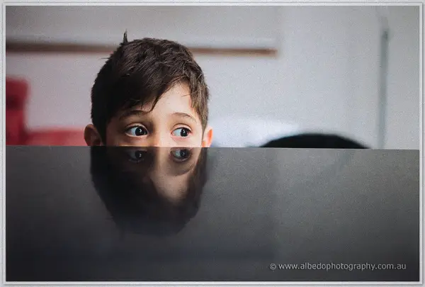 Young page boy peeks wide-eyed over a dark piano lid, his face reflected in the glossy surface