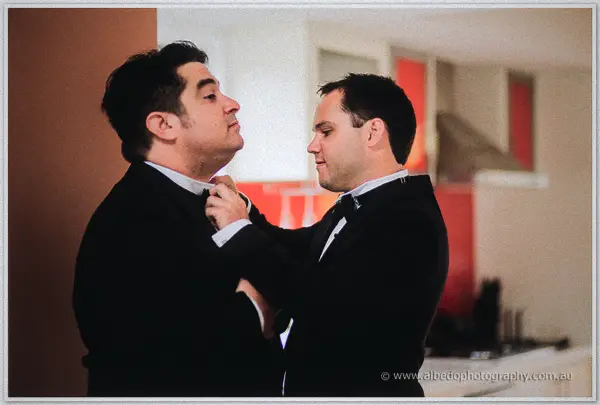 Groomsman straightens another's bow tie as they get ready in a bright modern apartment kitchen