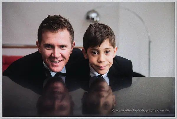 Groom and page boy grin side by side, chins resting on the polished piano lid