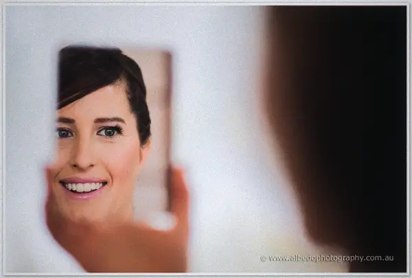 Bride beams at her reflection in a hand mirror as soft light fills the bright room