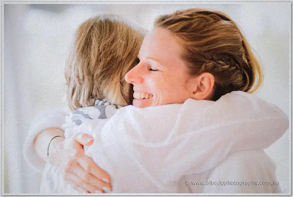 Two women in white robes embrace tightly, one laughing with eyes closed in the bright room